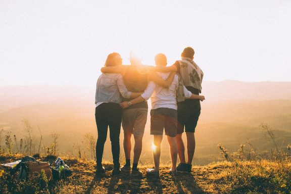 Eine Gruppe von vier Personen steht umarmt vor einem Sonnenuntergang in der Natur.
